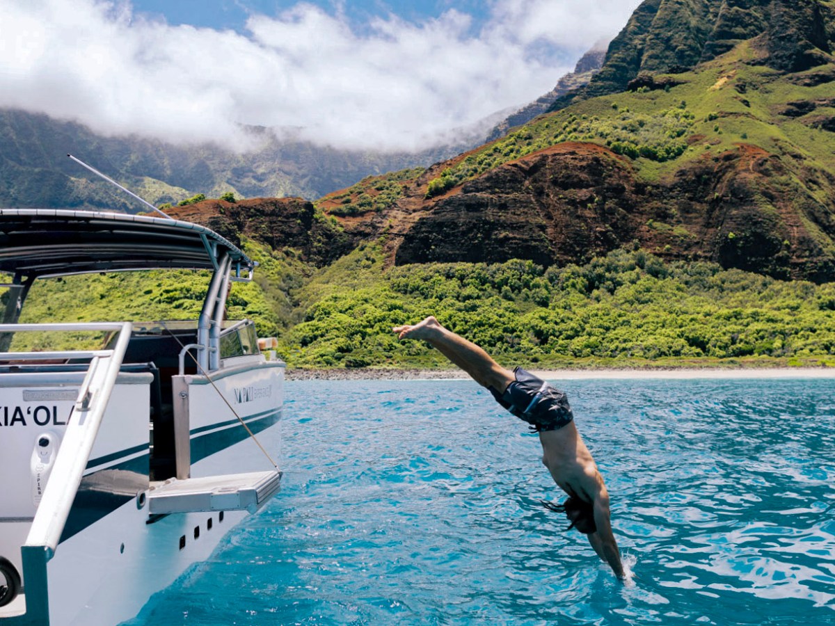 Person diving off boat into blue water near green mountains under a partly cloudy sky.
