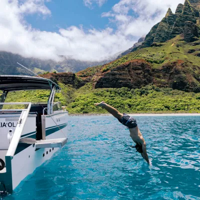 Person diving off boat into blue water near green mountains under a partly cloudy sky.