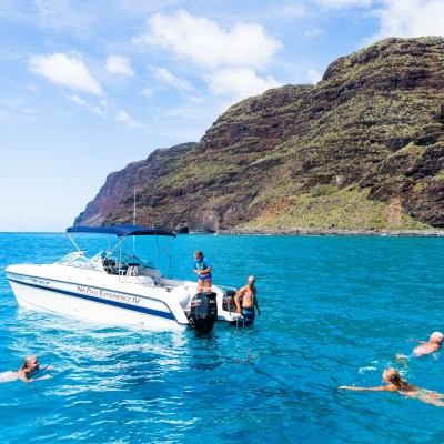 People swimming around a boat near rocky cliffs in clear blue water under a partly cloudy sky.