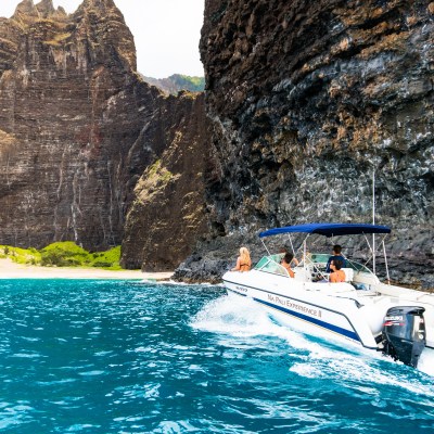 Boat with canopy cruises by rugged coastal cliffs and turquoise water, small beach visible.