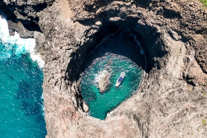 Aerial view of a boat in a circular, rocky sea cave with turquoise water, adjoining the ocean.