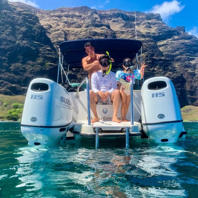 Three people on boat steps in snorkeling gear, cliffs and water in background.