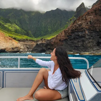 Woman sitting on a boat, looking at lush green cliffs under a cloudy sky.