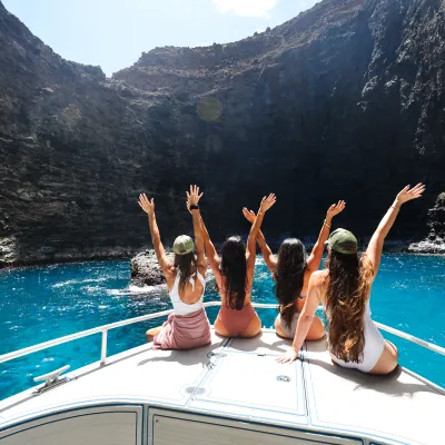 Four people on a boat with arms raised, surrounded by rocky cliffs and blue water.
