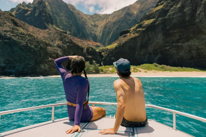 Two people on a boat admire a mountainous coastline and clear blue water.