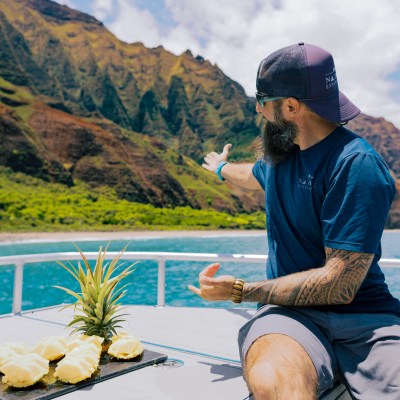 Man on boat gestures to scenic mountain, sliced pineapple beside him.