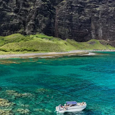 Boat with people in turquoise water near rocky cliffs and green hillside under blue sky.