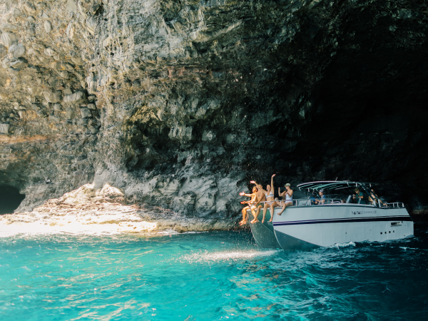 People jumping off a boat into blue water inside a large cave with rocky walls.
