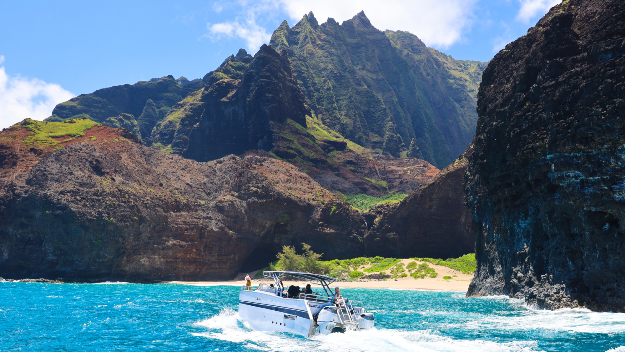 Boat sailing near rugged cliffs and a sandy beach with blue ocean and sunny sky.