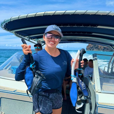 Person smiling on a boat holding snorkel gear, wearing a cap and sunglasses.