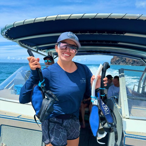 Person smiling on a boat holding snorkel gear, wearing a cap and sunglasses.