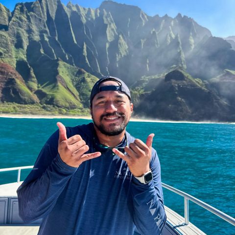 Person making shaka sign on a boat with stunning mountain backdrop and clear blue sky.