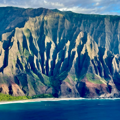 Dramatic coastal cliffs with jagged peaks and lush greenery above blue ocean waters under a cloudy sky.