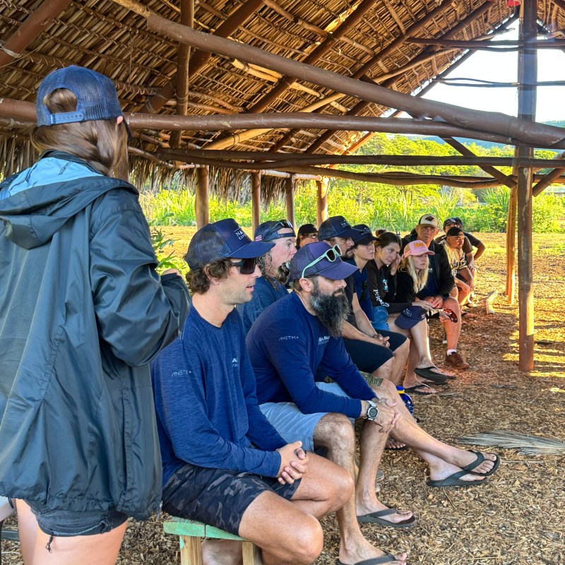 Group of people sitting under a thatched-roof shelter, wearing caps, surrounded by nature.