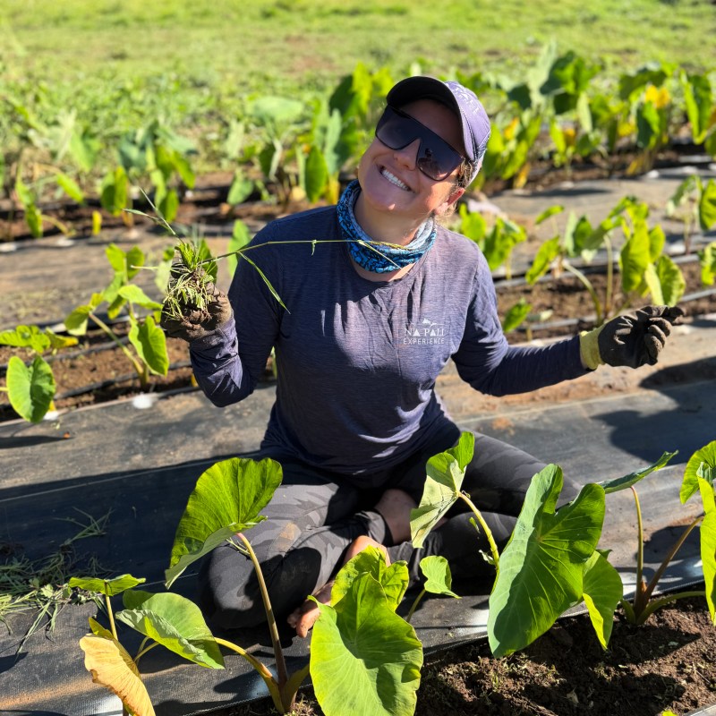 Smiling person in a cap and sunglasses sitting in a field with green plants, holding small weeds.