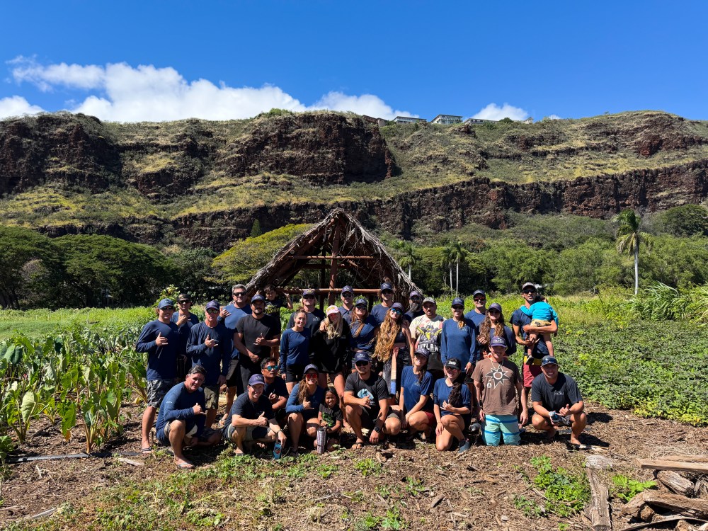 Group of people posing in front of a rustic hut with rocky hills in the background.
