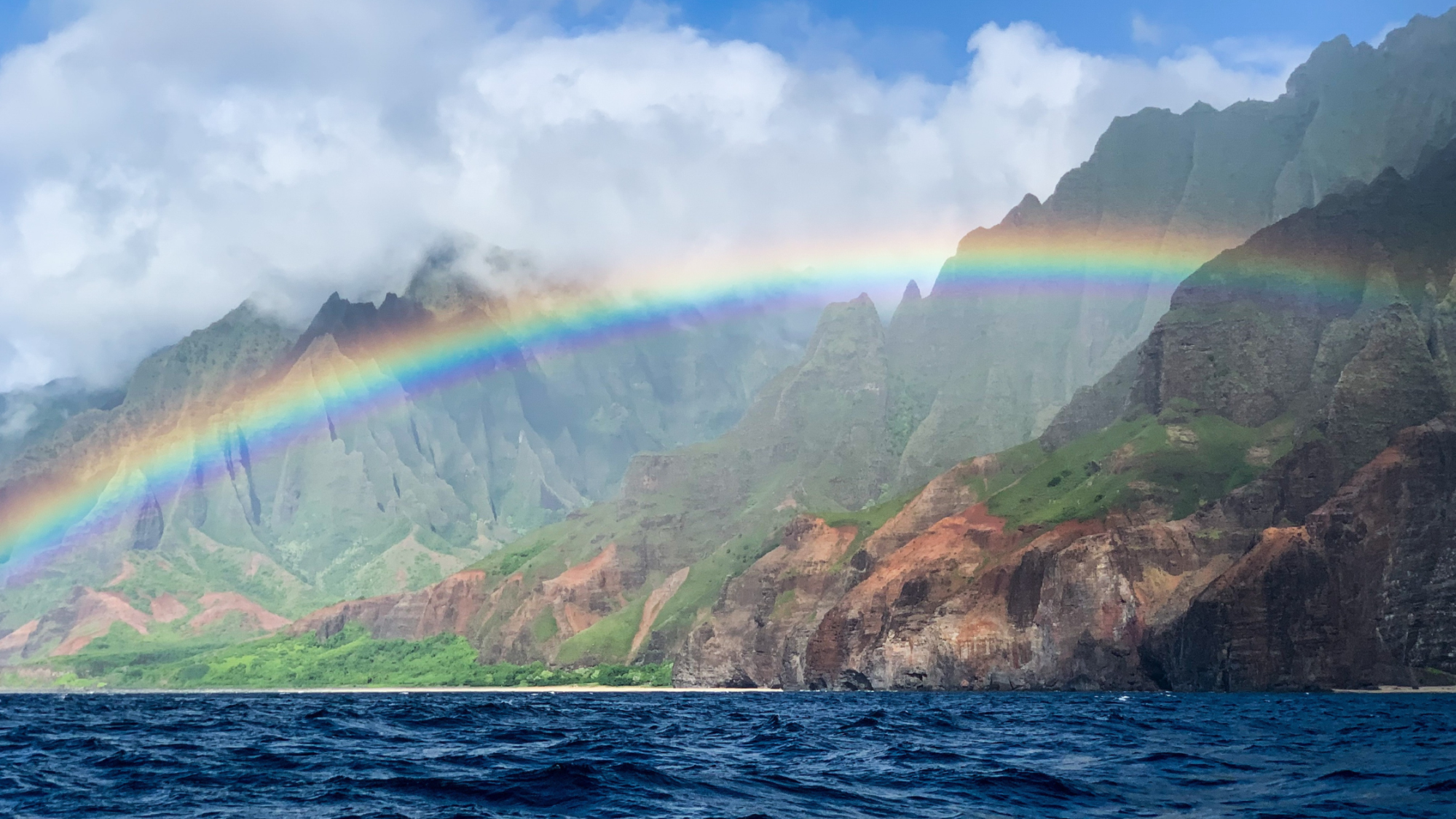 Rainbow arching over rugged coastal mountains with a cloudy sky.