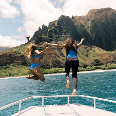 Two women jumping off a boat into the ocean with scenic mountains in the background.