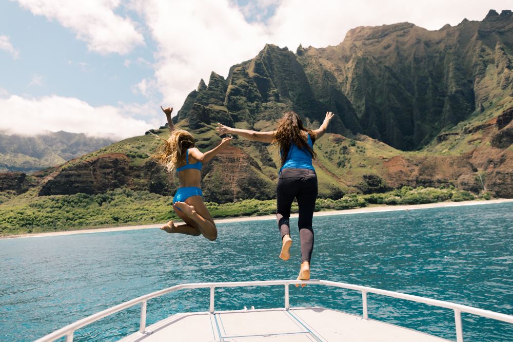 Two women jumping off a boat into the ocean with scenic mountains in the background.