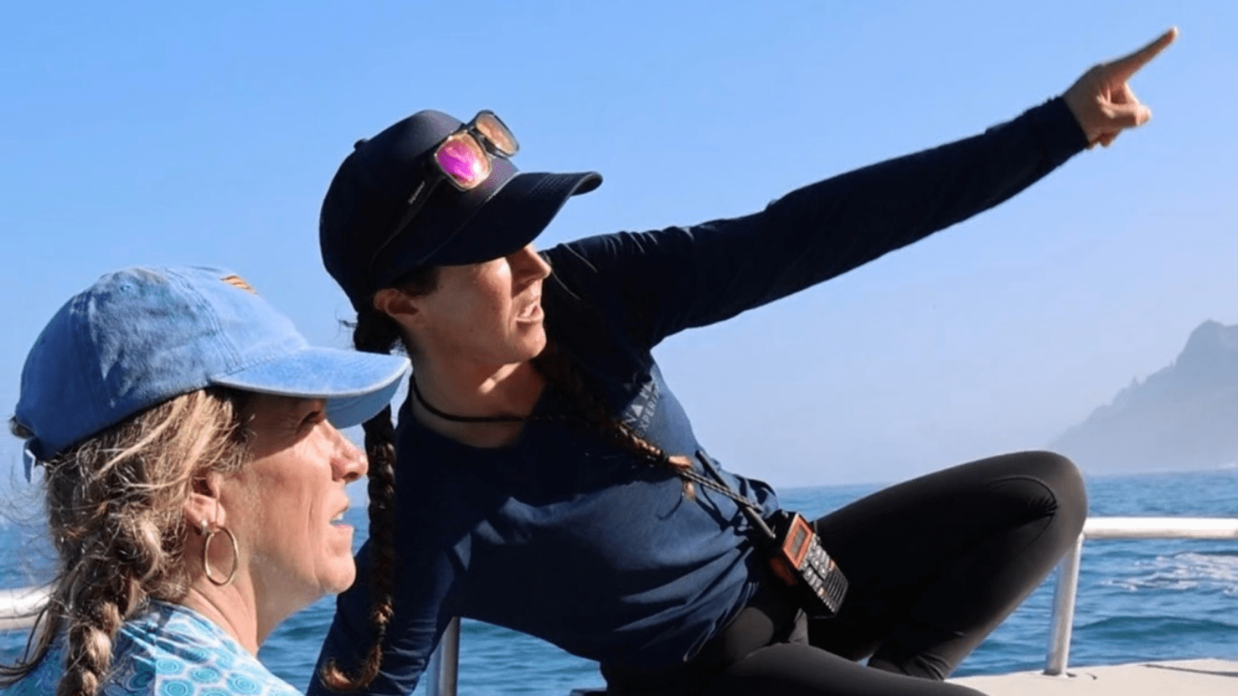 Two people in hats on a boat, one pointing towards distant mountains over the water.