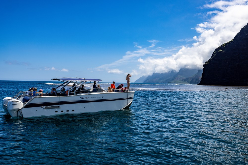 A tour boat with passengers on clear blue water near a mountainous coastline under a sunny sky.